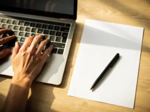 Handen typen op laptop toetsenbord naast wit papier en pen op houten bureau in warm natuurlijk licht